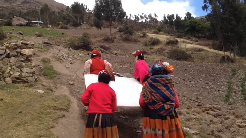 four women dressed in traditional brightly colored garb working together to carry a large sheet of drywall up a steep hill